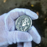 Silver coin held by a gloved hand against a blurred natural background
