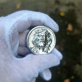 Silver coin with a face design held by a gloved hand against a blurred natural background